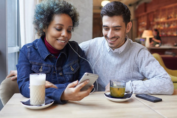 Obraz na płótnie Canvas Shot of interracial lovers sitting in a cafe enjoying their date, drinking coffee and tea and sharing audio tracks they listen together from cellphone, smiling and experiencing positive emotions.