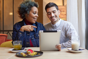 Indoor portrait of two young lovers spending their date together in cafe drinking coffee and tea and watching media on tablet PC that female is pointing to being interested in man’s opinion.