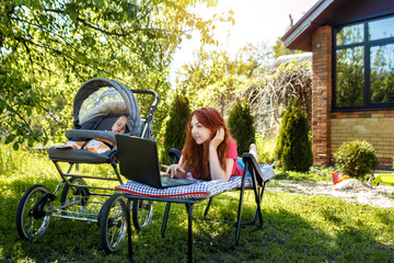 Young woman with her newborn baby lying on the soft armchair with laptop. Summer day. Mother with child outdoors. Motherhood.