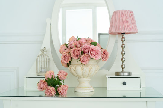 Bouquet of pink rores in vase and pink lamp on white boudoir table with oval mirror. Detail of the interior of the room with female boudoir