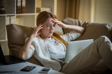 Attractive businesswoman on a couch with two cellphones and laptops. Exhausted woman in office late at night. Responsible executive working, having headache, hands on temples