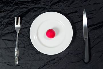 One red radish on white plate on black background with fork and knife.