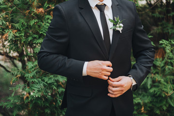 The groom in a suit and tie is standing on the background of greenery, on the bachelor's jacket is a boutonniere of flowers