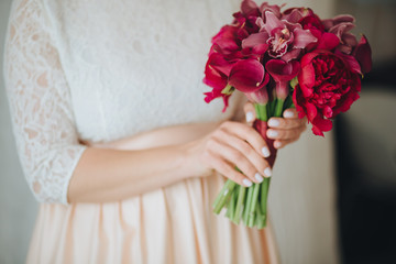 The bride in dress is standing by the window and holding a wedding bouquet of red flowers with a ribbon