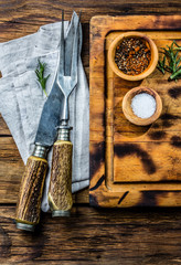 Cutting wooden board, cutlery set, rustic linen napkin, and spices on wooden background