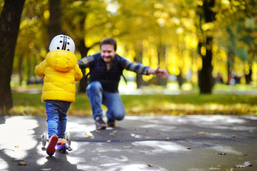 Middle age father showing his toddler son how to ride a scooter in a autumn park