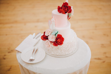 Three-tiered wedding cake is decorated with flowers and stands on the table next to a plate and cutlery