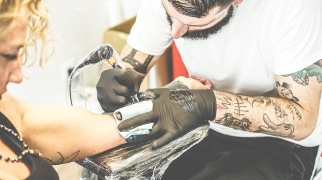 Bearded tattoo artist making tattoo inside ink studio on young blond woman