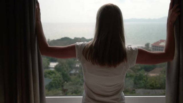 Woman in white t-shirt unveiling curtains and looking out of window. Enjoying the sea view outside