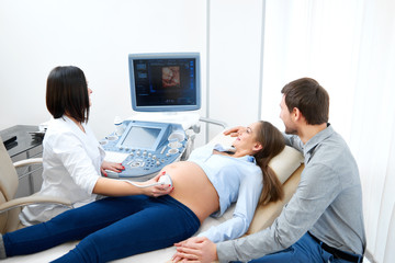 Happy loving pregnant couple during sonogram research at the fertility clinic. Female doctor performing ultrasound scanning for her pregnant patient sonogram medicine modern technology equipment.