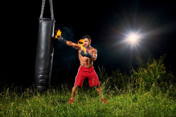 Shirtless muscular male boxer training outdoors at night exercising on a punching bag with his...