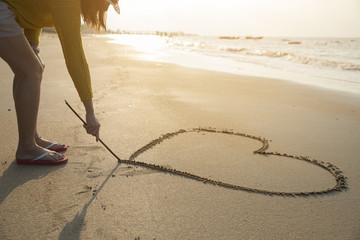 Beautiful woman writing message (heart symbol) on sand beach. - Powered by Adobe
