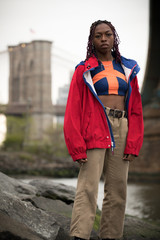 A young, black woman poses in front of NYC's Brooklyn Bridge. Dressed fashionably on a cloudy Spring day.