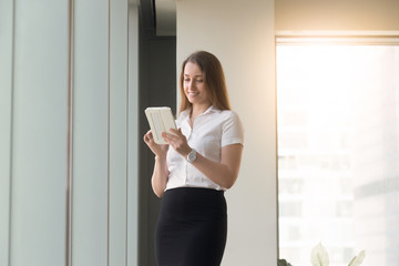 Smiling woman standing with computer tablet in hands. Happy female reading Internet news, entertains with mobile gaming, using app, write social network post. Successful businesswoman banking online
