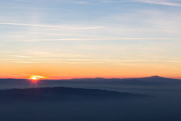 Sun coming down behind some misty mountains, the sky is orange and blue, with many long and thin clouds and jet trails