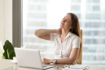Businesswoman feeling pain in neck after sitting at the table with laptop. Tired female suffering of office syndrome because of long hours computer work. Pretty girl massaging her tense neck muscles