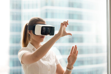 Beautiful girl using virtual reality glasses near bright window with skyscraper view outside....