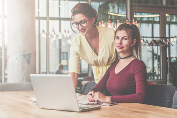 First girl is standing near table,second is sitting next to her.Teamwork.Girls blogging,working,learning online.