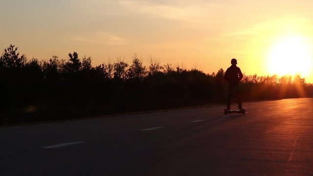 A boy on a gyroscooter rides into the sunset on the road near the forest.