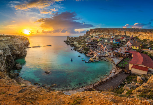 Il-Mellieha, Malta - Skyline view of the famous Popeye Village at Anchor Bay at sunset with traditional Luzzu boats, amazing colorful clouds and sky