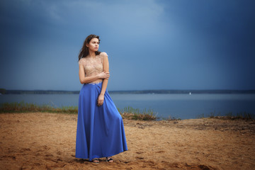 Fototapeta na wymiar Calm lonely fashion model walking on the sand in a cloudy day. Romantic, gentle, mystical, image of a girl in long blue skirt and lace blouse.