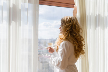 Beauty girl drinking coffee. Beautiful woman opening curtains, looking out the window and enjoying her morning coffee