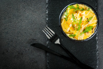 Casserole. French cuisine. Homemade potato gratin in a bowl for baking. On a black stone background. With leaves of fresh arugula for decoration. Top view copy space