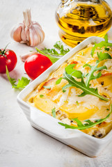 Casserole. French cuisine. Homemade potato gratin in a ceramic frying pan for baking. On a white marble stone background. With leaves of fresh arugula, tomatoes, olive oil.  copy space