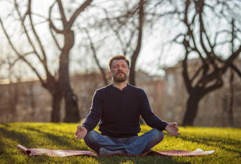 Mature man sitting on yoga mat in comfortable asana relaxing in park. Meditation in city life conditions