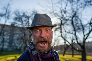 A bearded man in a hat screams in surprise. He's walking outside in the park.