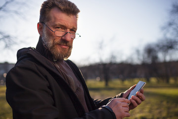 Obraz premium Portrait of mature man with beard in park. He hold phone in hands