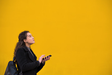 An Asian girl in a black coat and a phone with her hand walks down the street in front of a yellow wall - Powered by Adobe