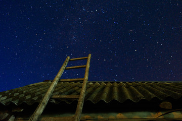Village. Old wooden ladder leaning against the barn with a slate roof in the night star sky. The staircase leads to the sky.