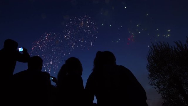 Silhouette of people who watch the fireworks in the night sky. Bright and colorful lights in the sky. A beautiful sight is watched by the girls. Red, green and orange lights in the blue sky.