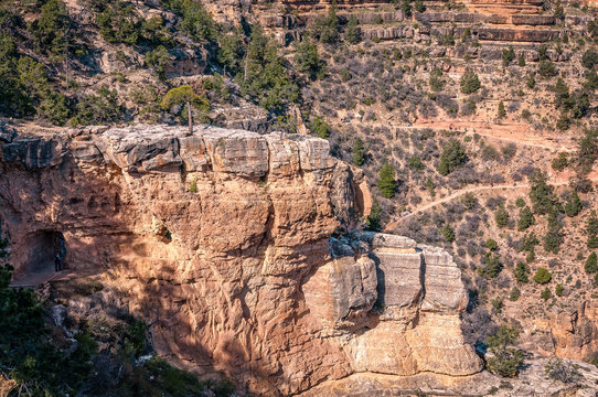 Interesting rock formation with pine trees on top. In the background, people are walking along the Bright Angel Trail on the South Rim of Grand Canyon National Park, Arizona, USA.