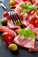Dried meat, jamon with cherry tomatoes, basil and olives on a black background. Selective focus.Top view. Copy space.