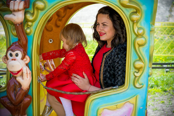 Obraz na płótnie Canvas A stylish mother in a red dress and a black jacket and a little daughter in a leather jacket for a walk in the city in an amusement park. The family is having fun and experiencing life