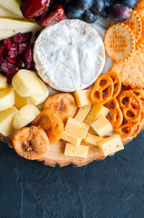 Cheese plate grape salted craker cookies dry dates cranberry fig pear pretzels on a wooden board on a black slate background.