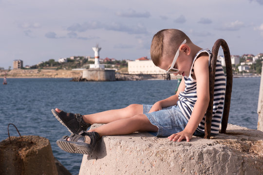 funny little boy in sunglasses and a vest sitting on breakwater on the sea background
