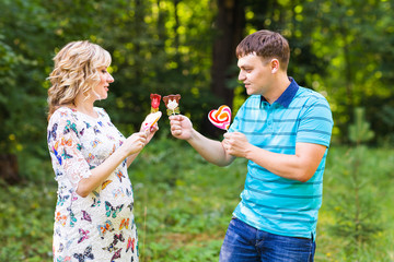 Fototapeta premium Pregnancy, family, happiness and fun concept - Man and pregnant woman have fun with candy in the park