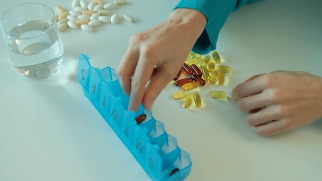 Woman sorts nutritional vitamins in blue tablet box on the week. Female adds one dark pill and three light pills in each cells of container by her hands. Designations of the days of the week is