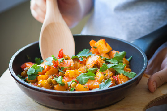 Woman holds a frying pan with vegetable stew on a cutting board and stir spoon. Healthy eating.