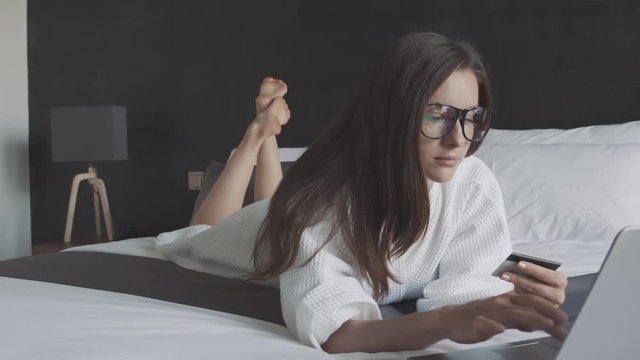 Woman in white bathrobe and glasses bue ticket with credit card and laptop, lie on the bed.