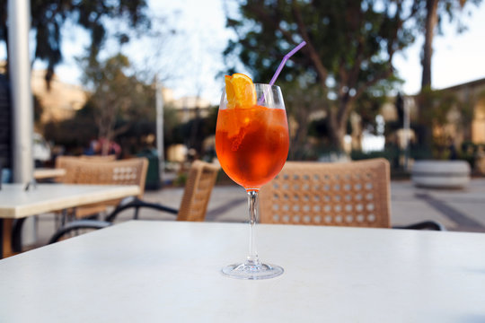 Aperol Spritz cocktail. Cocktail glass with an orange wedge and drinking straw on a table in cafe/restaurant outdoors. Traditional Italian aperitif made of prosecco and liqueur Campari, Cynar, Select