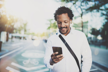 Happy american African man using smartphone outdoor.Portrait of young black cheerful man texting a sms message while listining to music.Blurred background, horizontal.Flare effects.