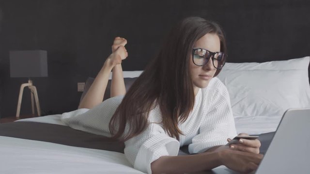 Woman in white bathrobe and glasses bue ticket with credit card and laptop, lie on the bed. Shopping online.