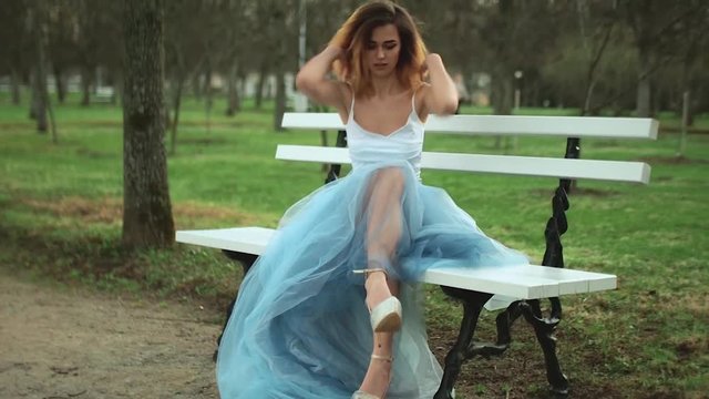 Attractive girl in silver and blue dress sits on bench in parkway buckles her high heeled shoes adjusts her tank top during photo shoot.