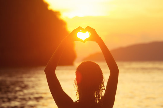 Heart shape making of female hands against bright sea sunset and sunny islands on horizon line