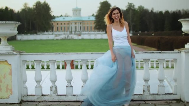 Attractive skinny girl in white and blue dress stands near white stone balustrade lauging and looking at camera during photo shoot.