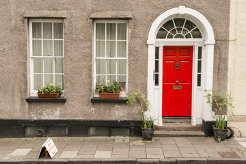 Facade of British traditional house with grey walls, red front door and two windows with flowers on...